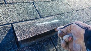 A roofing inspector examining asphalt shingles on a pitched residential roof with a clipboard in hand.