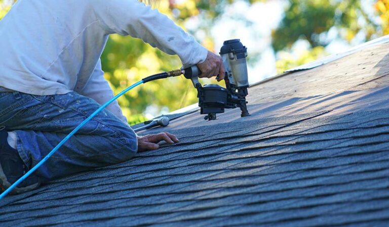A person on a ladder cleaning leaves from a house gutter with tree branches overhead.